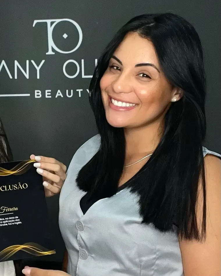Thuany Oliveira sorrindo diante do letreiro do estúdio, segurando um certificado de conclusão de curso